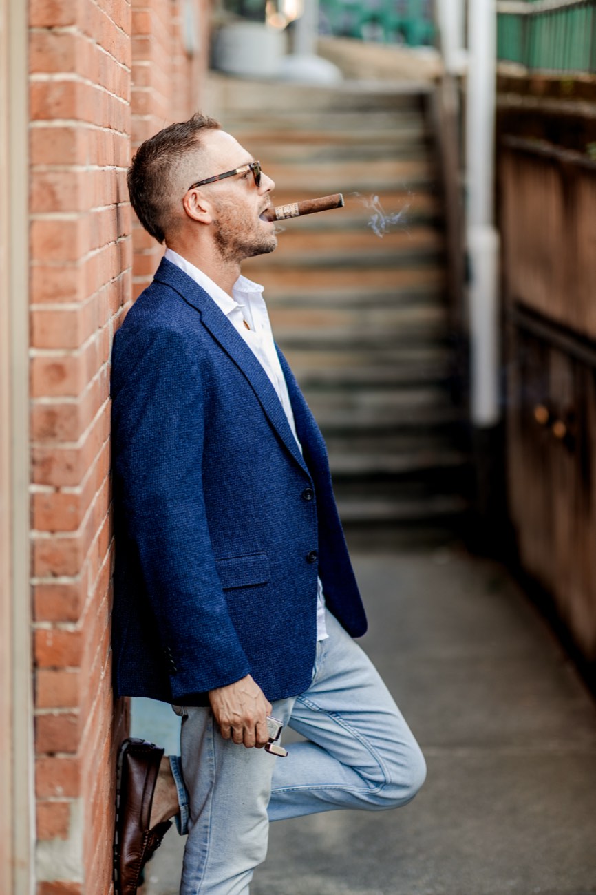 David leaning on a brick wall in an alley with a cigar.