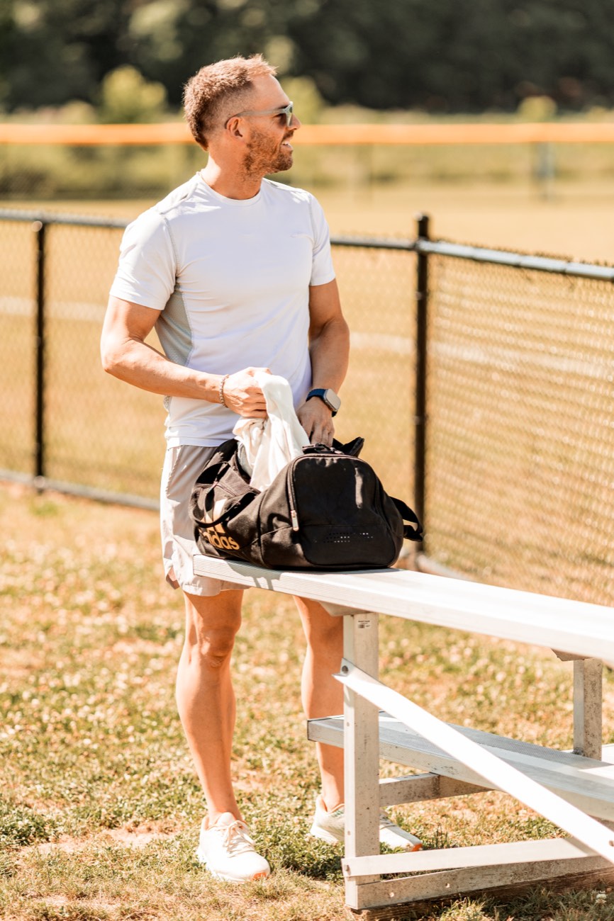 David carrying a gym bag past bleachers on a sunlit field.